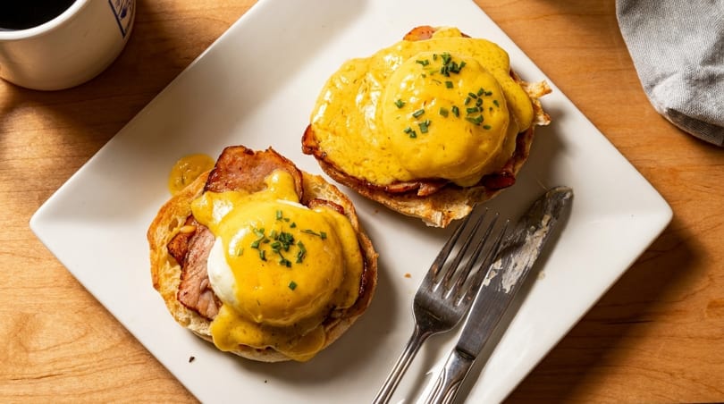 Two eggs Benedict with hollandaise sauce and chives on a white plate with cutlery.